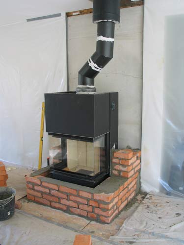 En modern svart öppen spis med glasfront omsluts delvis av röda tegelstenar och ovanför den sitter ett vinklat skorstensrör i svart metall. Det omgivande området är under uppbyggnad och väggarna är täckta med plastskivor.