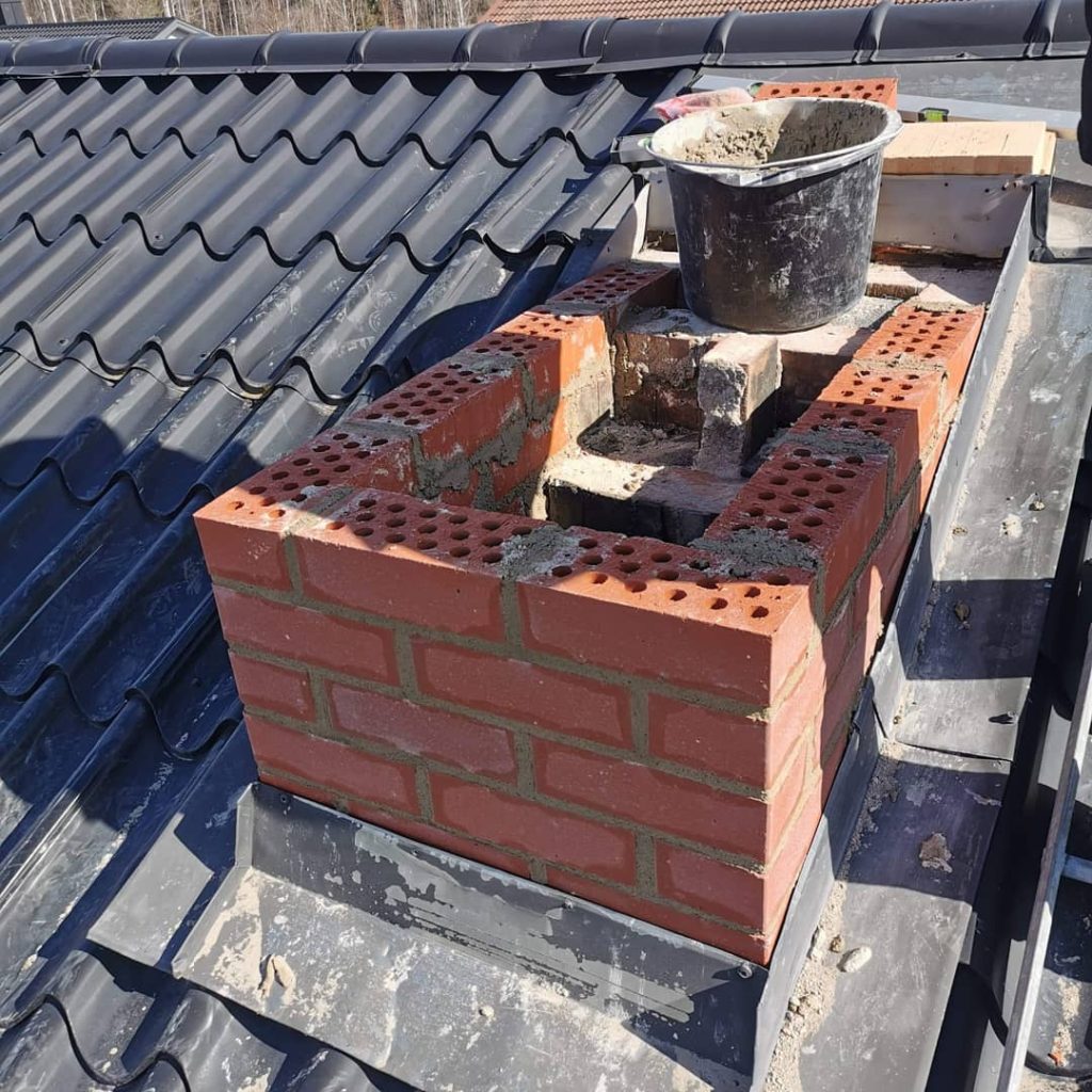 En delvis byggd tegelskorsten på ett tegeltak, med en hink med murbruk och verktyg placerad ovanpå. Skorstenens konstruktion är under uppbyggnad och visar exponerade tegelstenar och murbruksfogar.