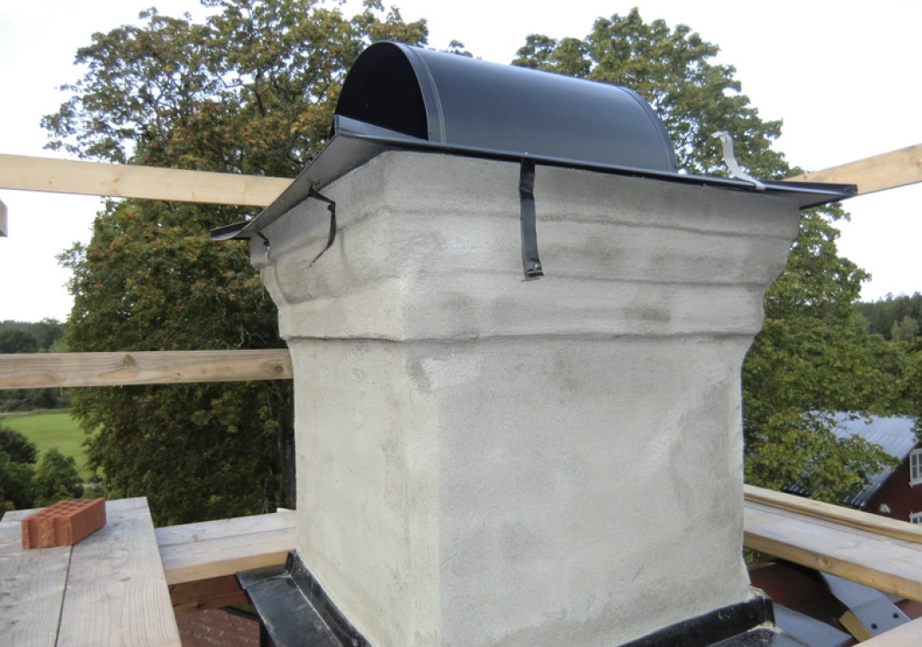 Närbild på en putsad skorsten med svart skorstenshatt i metall, omgiven av träställningar, med träd och en byggnad i bakgrunden.