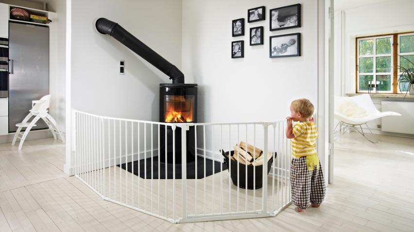 Ett litet barn står vid en vit säkerhetsgrind som omger en vedspis där det brinner. Veden ligger i en svart korg i närheten. Rummet är ljust med vita väggar, trägolv och inramade foton på väggen.