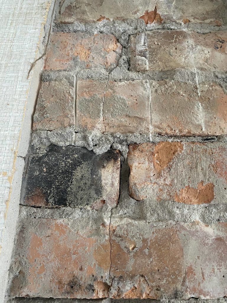 Besiktning Skorsten: Så går ett täthetstest till (Pris & Krav) 2 Närbild av en väderbiten tegelvägg i olika nyanser av rött, grått och svart, med sprickor, flisigt murbruk och strukturerade ytor. Till vänster syns kanten av ett mönstrat parti.