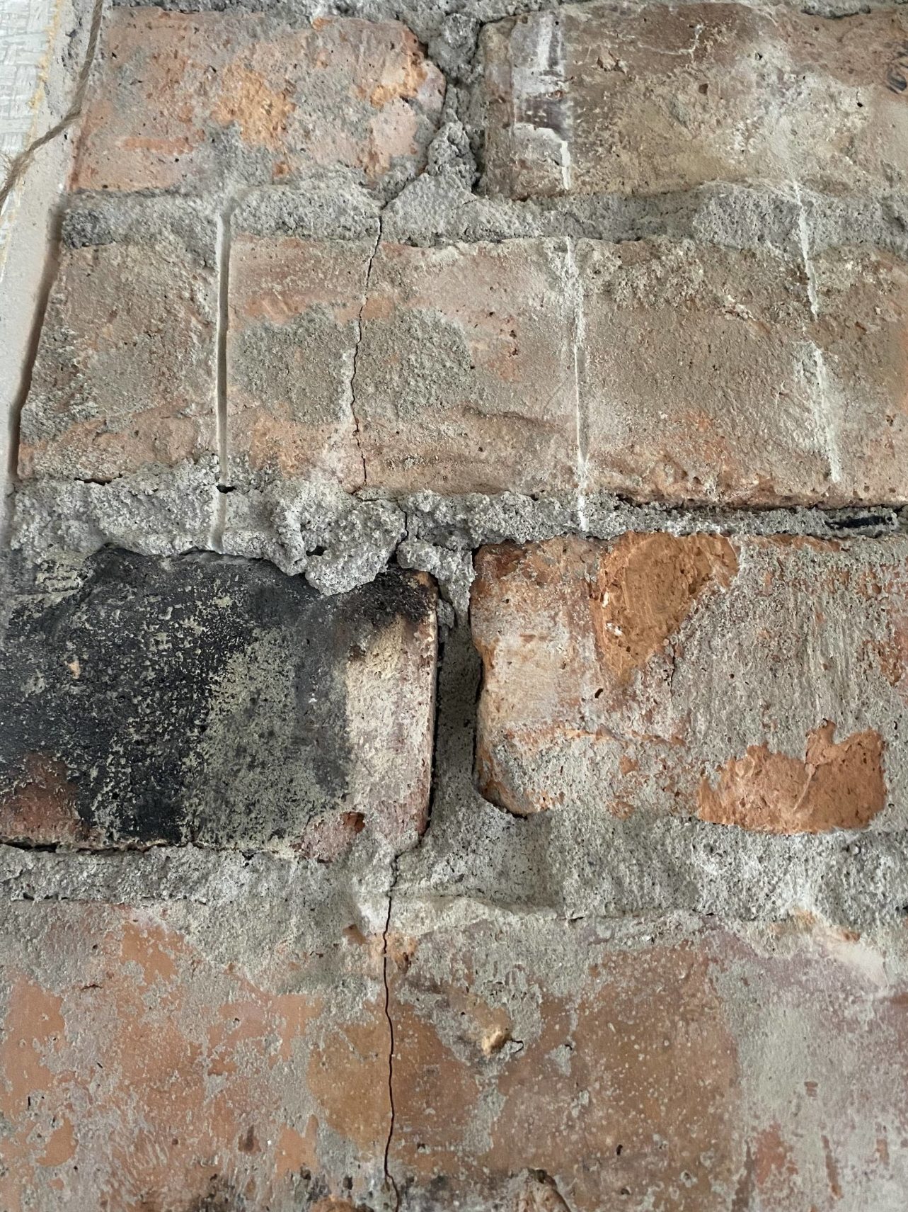 Närbild av en väderbiten tegelvägg i olika nyanser av rött, grått och svart, med sprickor, flisigt murbruk och strukturerade ytor. Till vänster syns kanten av ett mönstrat parti.