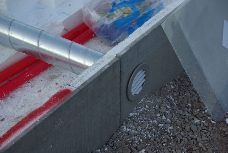 Tilluft Kamin: En komplett guide (Regler & Lösningar) 3 Närbild av en husgrund med skumisolering, en metallventil för tilluft kamin, röda rör, metallkanaler och plastfolie, omgiven av byggnadsavfall och grus.