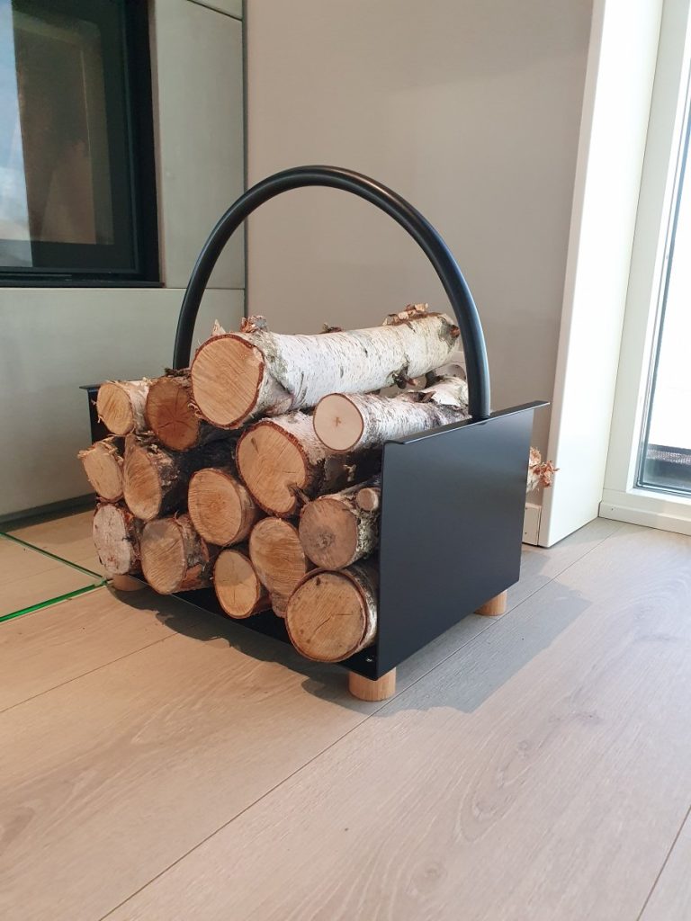 En svart vedhållare i metall med ett böjt handtag, fylld med staplade björkstockar, står på ett ljust trägolv nära ett fönster och en öppen spis.