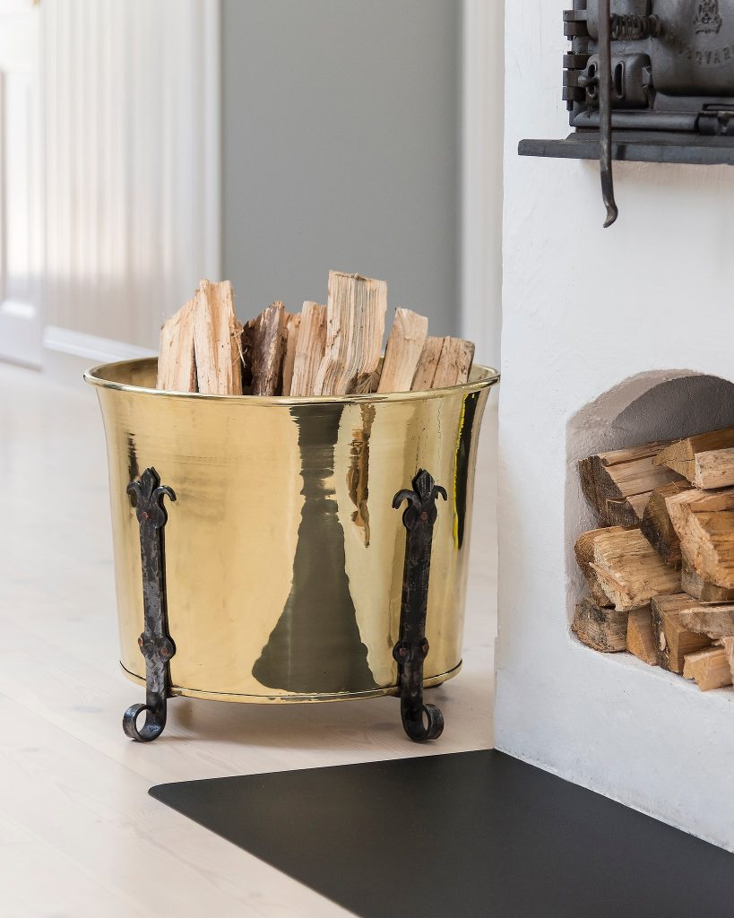 En vedhållare i blank mässing med svarta ben är fylld med huggen ved och placerad bredvid en vit öppen spis med staplade vedträn inuti.